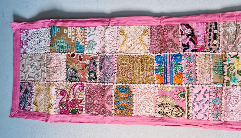 May include: A pink patchwork table runner with embroidered floral designs and beaded accents. The runner is made of various fabrics in shades of pink, beige, and brown.
