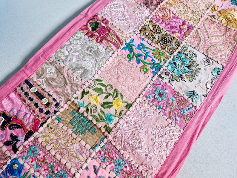 May include: A pink patchwork table runner with a variety of floral and geometric embroidery designs. The runner is made of different fabrics and textures, creating a unique and colorful look.