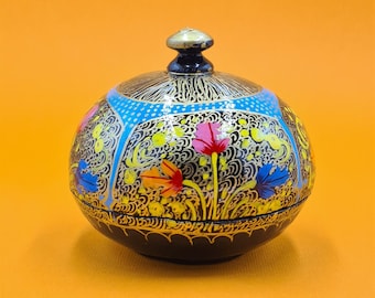 Può includere: Una scatola rotonda in legno dipinta a mano, nera, con un coperchio. La scatola è decorata con fiori colorati e motivi intricati in oro e blu.