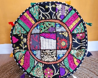 May include: Circular patchwork floor cushion with a black base and a vibrant, detailed design. The cushion incorporates various fabrics in pink, green, gold, and purple, with decorative tassels around the perimeter.