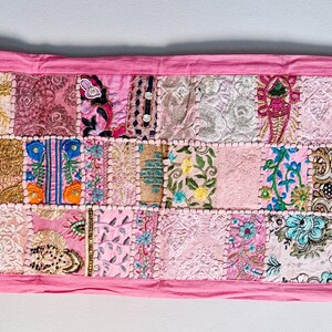 May include: A pink patchwork table runner with a variety of colorful floral and geometric patterns. The runner is made of different fabrics and is sewn together in a patchwork style.
