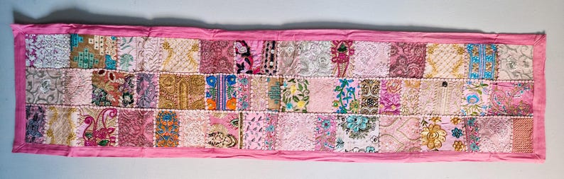 May include: A pink patchwork table runner with a variety of colorful floral and geometric patterns. The runner is made of different fabrics and is sewn together in a patchwork style.