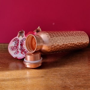 May include: A copper water bottle with a hammered finish and a matching lid. The bottle is lying on a wooden surface next to a half of a pomegranate.