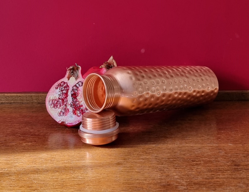 May include: A copper water bottle with a hammered finish and a matching lid. The bottle is lying on a wooden surface next to a half of a pomegranate.
