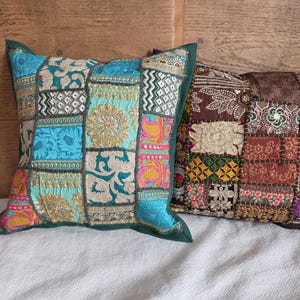 May include: Two square patchwork cushions. One is predominantly teal with gold and pink accents, the other is brown and red. The cushions are placed on a white surface, with a wooden background visible behind them.