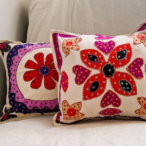 Può includere: Due cuscini decorativi con ricami floreali. Il cuscino più grande ha un motivo floreale rosso, rosa e nero su sfondo bianco. Il cuscino più piccolo ha un motivo floreale rosso, rosa e viola su sfondo bianco.