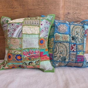 Puede incluir: Dos cojines cuadrados con un diseño de patchwork hecho de paneles de tela bordada. El cojín de la izquierda muestra patrones florales en verde claro, naranja y blanco. El de la derecha presenta tonos azules, dorados y verde azulado con detalles en hilo metálico.