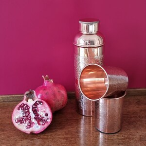 K&ouml;nnte beinhalten: Zwei Kupferbecher und ein Kupfer-Cocktail-Shaker mit einem gravierten Design. Der Shaker hat einen silbernen Deckel. Zwei Granat&auml;pfel, einer ganz und einer halbiert, befinden sich im Vordergrund.