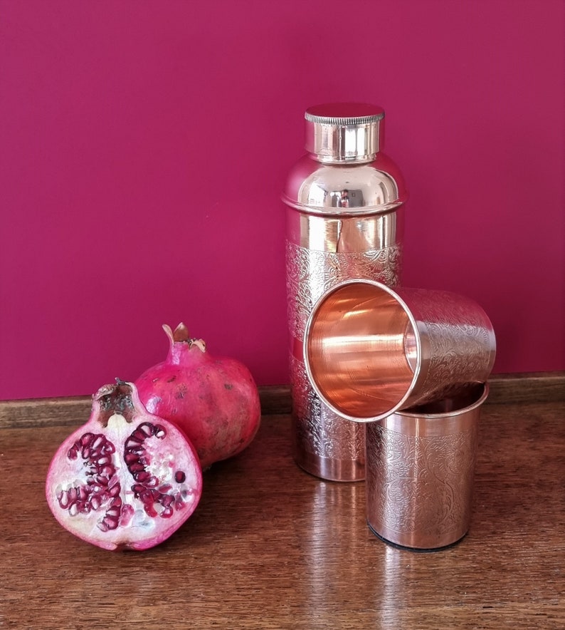 K&ouml;nnte beinhalten: Zwei Kupferbecher und ein Kupfer-Cocktail-Shaker mit einem gravierten Design. Der Shaker hat einen silbernen Deckel. Zwei Granat&auml;pfel, einer ganz und einer halbiert, befinden sich im Vordergrund.