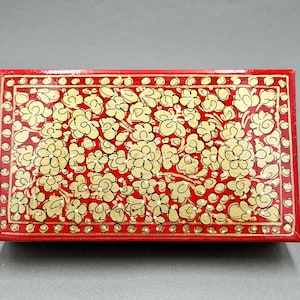 Puede incluir: Una caja roja con un diseño floral dorado. La caja está hecha de papel maché y tiene una tapa con bisagras.