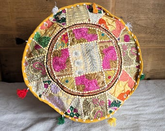 May include: A circular patchwork cushion in a variety of colours, including pink, gold, green, and yellow. The cushion has a central circular design and is trimmed with a yellow border and small tassels.