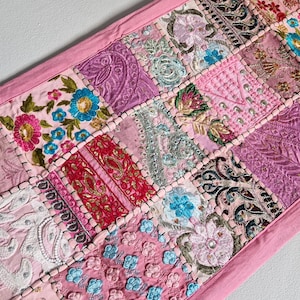 Może przedstawiać: Różowy bieżnik stołowy w patchworku z haftowanymi wzorami kwiatowymi i koralikowymi akcentami. Bieżnik wykonany jest z różnych tkanin i faktur, tworząc unikalny i kolorowy element.