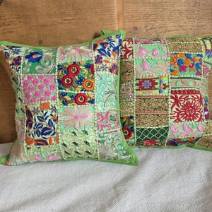 Può includere: Due cuscini decorativi con un design patchwork. I cuscini presentano una varietà di toppe ricamate colorate nelle tonalità del verde, rosa, rosso e blu. I cuscini hanno un bordo verde e sono posizionati su una superficie bianca.