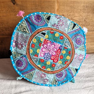 Può includere: Cuscino rotondo ricamato con un vivace motivo floreale. Il cuscino presenta uno sfondo turchese con toppe di vari motivi e colori, tra cui rosa, rosso, giallo e viola. Ha un bordo blu con cuciture bianche e piccole nappe.