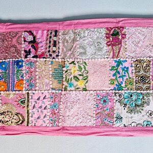 May include: A pink patchwork table runner with various floral and geometric embroidery designs. The runner is made of different fabrics and textures, creating a unique and colorful piece.