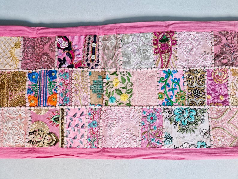 May include: A pink patchwork table runner with various floral and geometric embroidery designs. The runner is made of different fabrics and textures, creating a unique and colorful piece.
