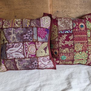 Puede incluir: Dos cojines decorativos con diseños de patchwork. Los cojines presentan una mezcla de telas burdeos, moradas y doradas con intrincados bordados y adornos. Los cojines tienen forma cuadrada y un borde burdeos liso.