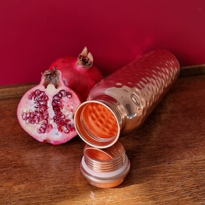 May include: A copper water bottle with a hammered finish and a matching lid. The bottle is open and the lid is sitting next to it. A half of a pomegranate is next to the bottle.