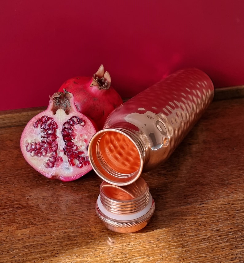 May include: A copper water bottle with a hammered finish and a matching lid. The bottle is open and the lid is sitting next to it. A half of a pomegranate is next to the bottle.