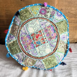 Può includere: Cuscino rotondo da pavimento patchwork con un design bohémien vibrante. Il cuscino presenta un mix di motivi floreali e paisley nei toni del verde, viola e blu. Ha un bordo turchese con nappe colorate e un bordo cucito decorativo.