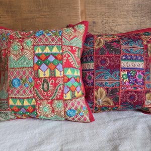 May include: Two decorative cushions with patchwork designs. The cushions have a red border and a vibrant mix of colours including green, blue, yellow, and purple. The patchwork includes floral and geometric patterns.