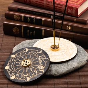 Könnte beinhalten: Zwei runde Räucherstäbchenhalter mit Himmelsdesigns. Einer ist schwarz mit goldenen Akzenten, der andere ist helles Holz. Beide haben einen goldfarbenen Metall-Räucherstäbchenhalter in der Mitte. Räucherstäbchen sind in die Halter eingesetzt.