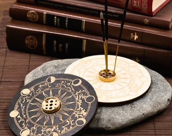 May include: Two round incense holders with celestial designs. One is black with gold accents, the other is light wood. Both have a gold-colored metal incense stick holder in the center. Incense sticks are inserted in the holders.