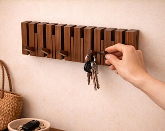 May include: A wooden key holder with a series of rectangular blocks and hooks. The key holder is mounted on a wall. A hand is reaching for a key. A woven basket and a small bowl with a remote and coins are on a wooden surface below.
