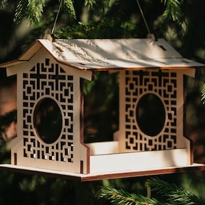 Op de afbeelding: Een houten vogelvoederhuisje met een decoratief geometrisch ontwerp. De voederbak heeft een dak en een platform waar vogels op kunnen zitten. De voederbak hangt aan een tak.