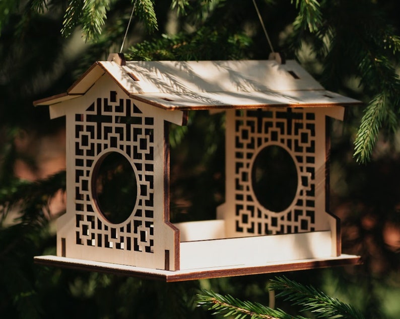 Op de afbeelding: Een houten vogelvoederhuisje met een decoratief geometrisch ontwerp. De voederbak heeft een dak en een platform waar vogels op kunnen zitten. De voederbak hangt aan een tak.