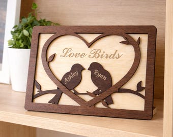 May include: A wooden plaque with a heart-shaped cutout and the words "Love Birds" engraved. Two bird silhouettes sit on a branch within the heart. The names "Ashley" and "Ryan" are engraved on the birds. The frame is dark brown.