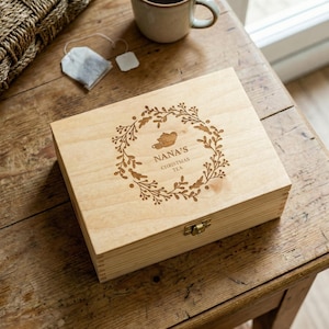 Peut inclure: Une boîte à thé en bois clair avec une couronne décorative et l'inscription "NANA'S CHRISTMAS TEA" gravée sur le couvercle. Un sachet de thé et une tasse de thé sont posés sur une table en bois.