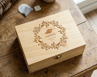 May include: A light-colored wooden tea box with a decorative wreath and the text "NANA'S CHRISTMAS TEA" engraved on the lid. A tea bag and a mug of tea are next to the box on a wooden table.
