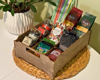 May include: A wooden gift box filled with various food items, including jars, packets, and a jar of jam. The box contains colorful straws and is placed on a woven mat. A potted plant is in the background.