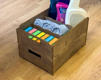 May include: A wooden storage box with compartments, holding cleaning supplies. The box is stained a dark brown and contains spray bottles, colorful sponges, and folded cloths. The box has a handle cut-out on the front.