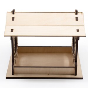 Op de afbeelding: Een houten vogelvoederhuisje met een schuin dak en een decoratief uitgesneden ontwerp. De voederbak is gemaakt van lichtbruin hout met donkerbruine accenten.