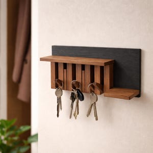 Può includere: Un portachiavi in legno con un supporto grigio scuro. Il portachiavi ha un piccolo ripiano e una serie di doghe verticali in legno per riporre le chiavi. Diversi mazzi di chiavi sono appesi al supporto. Il portachiavi è montato su una parete bianca.