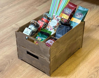 May include: A wooden organizer with multiple compartments filled with various items. The contents include tea bags, jars of snacks, colorful straws, and small packets. The organizer is made of light brown wood and sits on a wooden surface.