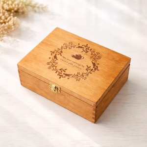 May include: A wooden tea box with a decorative wreath and teapot design engraved on the lid. The box is rectangular with a brass clasp. The text on the lid reads "May your mornings be treated with jasmine."