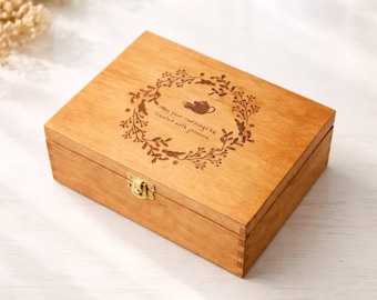 May include: A wooden tea box with a decorative wreath and teapot design engraved on the lid. The box is rectangular with a brass clasp. The text on the lid reads "May your mornings be treated with jasmine."