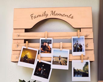 May include: A wooden photo display with the words "Family Moments" engraved on the top. The display holds several photos clipped to it with small wooden clothespins. The photos are of various sizes and feature different scenes.