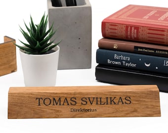 Op de afbeelding: Een houten bureau-naamplaatje met de tekst "TOMAS SVILIKAS Direktorius" gegraveerd in zwarte letters. Het naamplaatje ligt op een licht houten oppervlak.