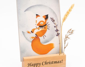 May include: A wooden card holder with the text "Happy Christmas!" engraved on the front. A card with a watercolor illustration of a fox sitting on a crescent moon is displayed in the holder. Dried flowers and wheat stalks are in the holder.