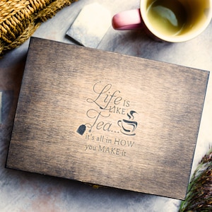 Può includere: Una scatola di legno con una macchia marrone scuro. Il coperchio ha una citazione incisa che recita "La vita è come il tè... tutto sta nel modo in cui lo fai".