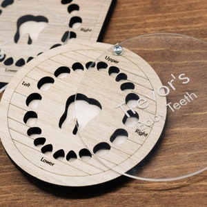 May include: A wooden baby teeth chart with a clear plastic overlay. The chart has 20 spaces for teeth, labelled "Upper", "Lower", "Left", and "Right". The overlay has the name "Trevor's Baby Teeth" printed on it.