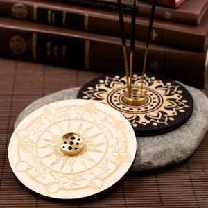 May include: Two wooden incense holders with intricate designs. One is white with a sun and moon motif, the other is black with a mandala pattern. Both have a brass center for burning incense.