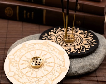 May include: Two wooden incense holders with intricate designs. One is white with a sun and moon motif, the other is black with a mandala pattern. Both have a brass center for burning incense.