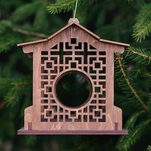 Op de afbeelding: Een houten vogelvoederhuisje in de vorm van een huis met een geometrisch patroon en een ronde opening. De voederbak hangt aan een tak.