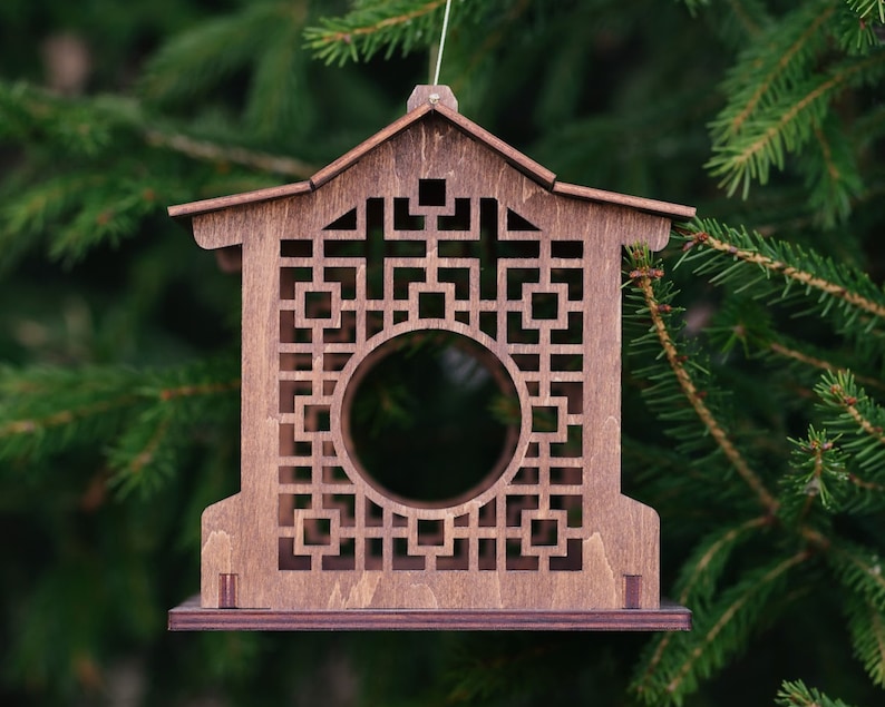 Op de afbeelding: Een houten vogelvoederhuisje in de vorm van een huis met een geometrisch patroon en een ronde opening. De voederbak hangt aan een tak.
