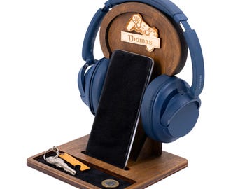 May include: A wooden headphone stand with a built-in phone holder and a small tray for keys and coins. The stand features a circular design with a carved detail and the name "Thomas". Blue headphones and a black smartphone are displayed.